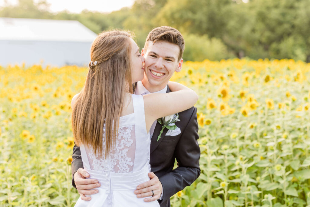 Bride kissing Groom's Cheek in a field of sunflowers on their wedding day.