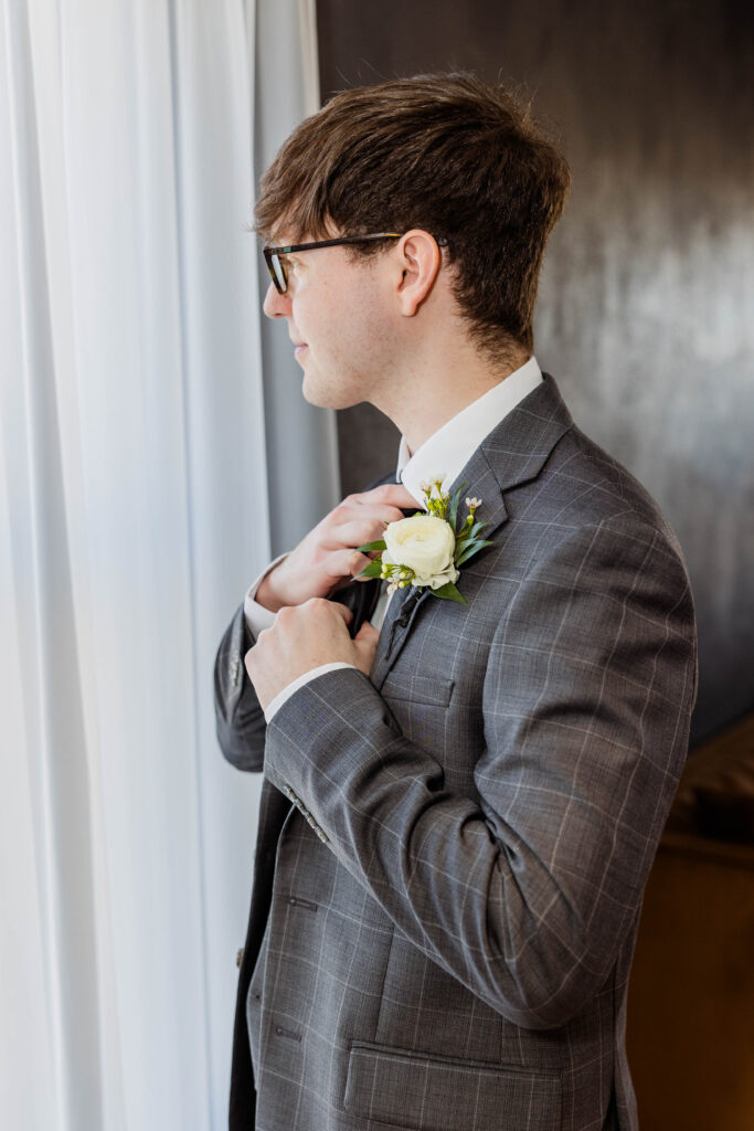 Groom adjusting his tie looking out the window on her wedding day.